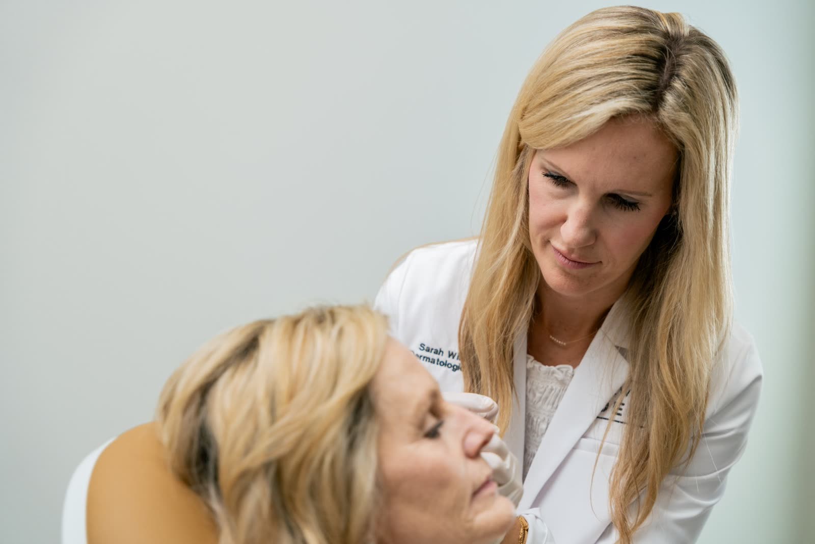 Sarah Wilson, MD, in her white coat, with a gloved hand at a reclining guest’s cheek during a filler treatment
